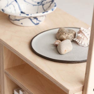 May include: A light wood shelf unit with three shelves. The top shelf has a white ceramic bowl with blue splatter paint, a grey ceramic plate with a dark grey rim, and three decorative stones and shells. The bottom shelf has two white books.