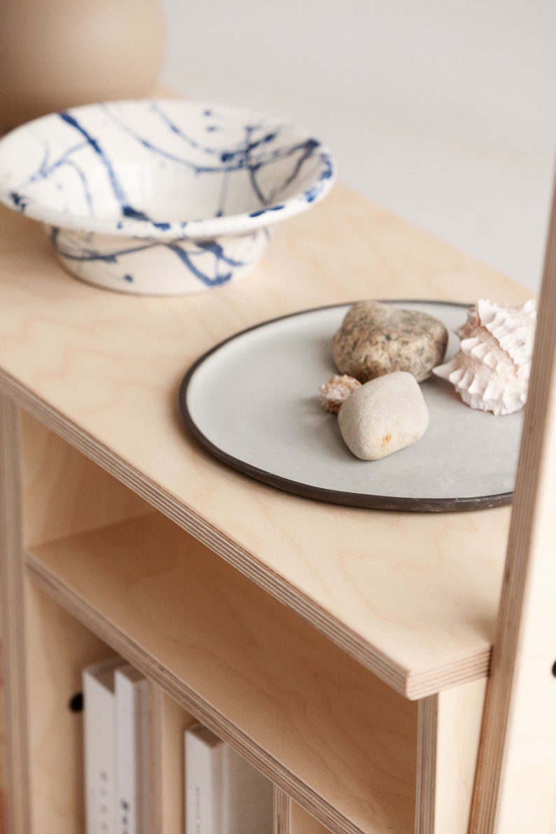 May include: A light wood shelf unit with three shelves. The top shelf has a white ceramic bowl with blue splatter paint, a grey ceramic plate with a dark grey rim, and three decorative stones and shells. The bottom shelf has two white books.