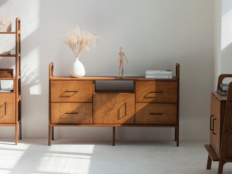 Op de afbeelding: Een houten dressoir met vier lades en een kastdeur. Het dressoir heeft een lichte houtkleur en zwarte metalen handgrepen. Er staat een klein houten beeldje en een stapel boeken op het dressoir.
