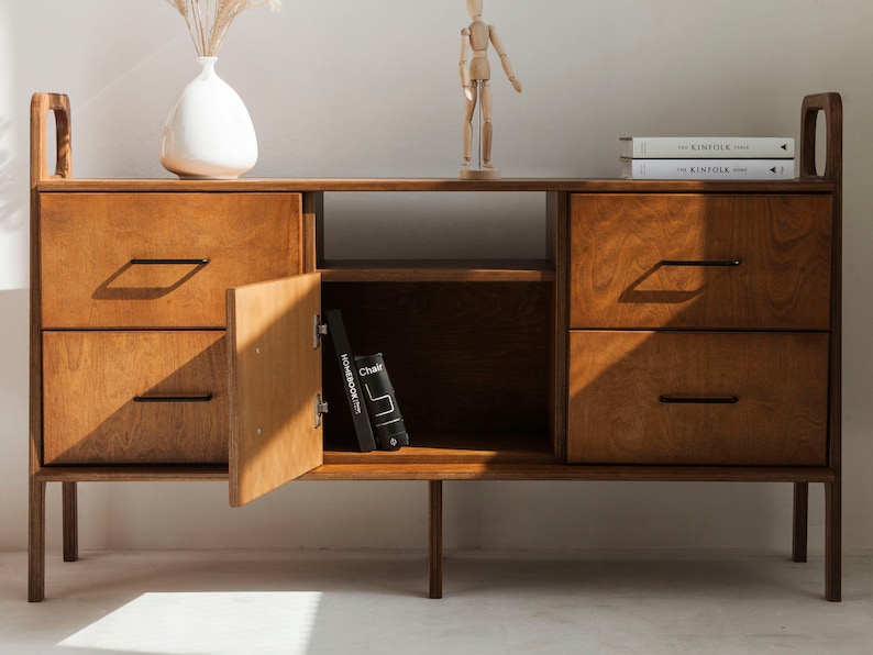 Op de afbeelding: Een houten kast met twee lades en een deur aan elke kant. De kast heeft een plank in het midden en staat op vier poten. De kast is gemaakt van lichtbruin hout en heeft zwarte metalen handgrepen.