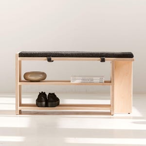 May include: A light-colored wooden bench featuring a dark charcoal gray upholstered seat cushion. The bench includes two open shelves for storage, with a small beige ceramic bowl, two stacked books, and a pair of black shoes placed on them.