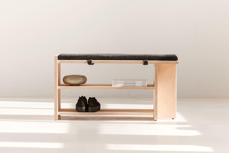 May include: A light-colored wooden bench featuring a dark charcoal gray upholstered seat cushion. The bench includes two open shelves for storage, with a small beige ceramic bowl, two stacked books, and a pair of black shoes placed on them.