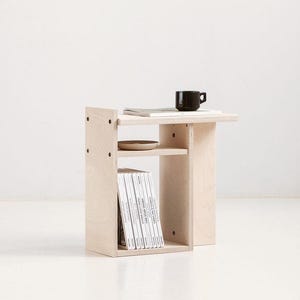 May include: A light-colored plywood side table featuring a flat top surface, a middle shelf, and a lower storage compartment. The table holds a black mug, a thin book, a small ceramic dish, and a row of vertical books on the bottom shelf.