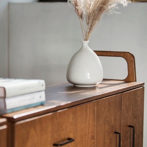 Op de afbeelding: Een houten kast met twee deuren en een lade. De kast heeft zwarte handgrepen en een lichte houtkleur. Een witte vaas met gedroogd pampasgras staat op de kast.