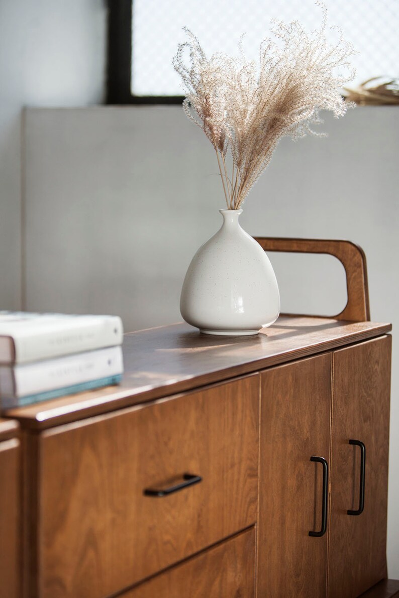 Op de afbeelding: Een houten kast met twee deuren en een lade. De kast heeft zwarte handgrepen en een lichte houtkleur. Een witte vaas met gedroogd pampasgras staat op de kast.
