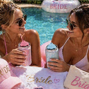 K&ouml;nnte beinhalten: Zwei Frauen in einem Pool halten glitzernde Getr&auml;nkedosen mit Deckeln. Rosa "Bride" Baseballkappe, Handtuch und Kosmetiktasche. Ein "Bride" Pool-Schwimmer ist im Hintergrund. Die Getr&auml;nkedosen haben einen "Codiup" Deckel.