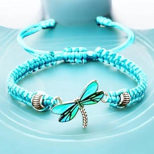 May include: A turquoise braided bracelet with a dragonfly charm. The dragonfly has turquoise wings with black details and a silver-colored body. The bracelet is adjustable and has silver-colored metal accents.