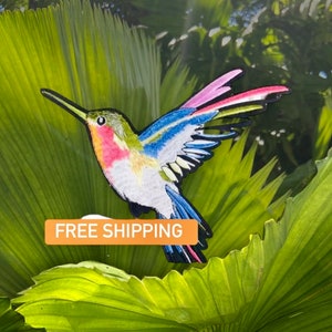 May include: A colorful hummingbird with blue, pink, and green feathers is perched on a green leaf. The hummingbird is in flight with its wings spread out. The background is a blurred image of green leaves.