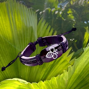 May include: A purple leather bracelet with a black braided cord and a silver bee charm. The bracelet is on a green leafy background.