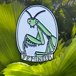 May include: A green praying mantis with a white background and the word "FEMINISM" written on a scroll below it. The pin is a metal enamel pin.