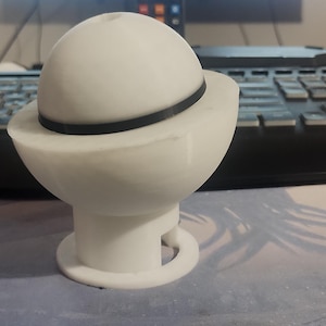 Puede incluir: Un objeto blanco impreso en 3D que se asemeja a un planeta con un anillo, apoyado sobre una base. La parte superior esférica tiene una banda negra alrededor de su centro. La base tiene una forma acampanada con un pie circular. El objeto está sobre un escritorio con un teclado en el fondo.