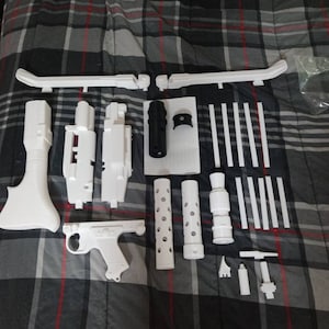 May include: A collection of white 3D-printed components, including long curved sections, cylinders with perforations, and a gun-shaped part. A black cylindrical piece and other small parts are also visible. The items are laid out on a plaid patterned surface.