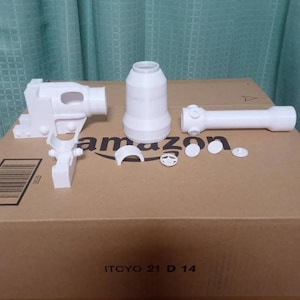 Pode incluir: Um conjunto de peças brancas impressas em 3D, incluindo uma estrutura semelhante a uma pistola, uma peça cilíndrica e um tubo longo com acessórios circulares. As peças estão dispostas numa caixa de cartão com o logótipo da Amazon e o texto "ITCYO 21 D 14".