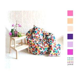 May include: A colorful beanbag chair with a geometric pattern. The chair is filled with colorful cubes and has a matching pillow. The chair is sitting on a white surface with a small wooden stool and a potted plant.