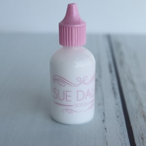 May include: A white bottle with a pink cap and the text "Sue Dale Designs" on the label.