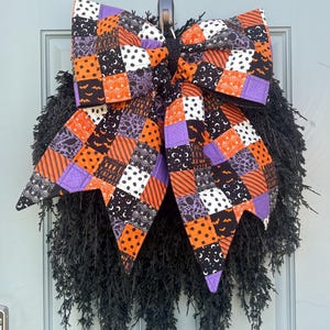 May include: A large decorative bow made of quilted fabric featuring a patchwork pattern of orange, purple, black, and white squares with various Halloween-themed prints, attached to a textured black wreath with long, wispy, needle-like foliage.