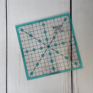 May include: A clear, square quilting ruler with a grid pattern and floral accents. The ruler has measurements in inches and features the text "Cute Cuts". The edges are a light teal color.