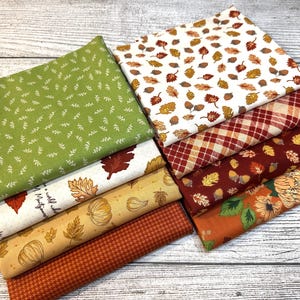 May include: A collection of folded fabric squares featuring autumn-themed patterns including green with white sprigs, white with colorful leaves and acorns, red and white plaid, dark red with leaves, and orange with pumpkins and floral motifs.