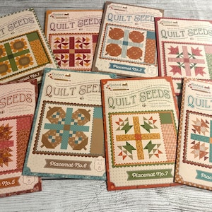 May include: A collection of eight rectangular paper quilt pattern booklets titled Quilt Seeds by Baked With Love. Each booklet features a unique colorful geometric quilt block design, specific placemat numbering, and a small bee illustration.