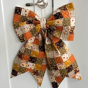 May include: A large decorative bow constructed from quilted fabric patches in shades of orange, brown, yellow, and cream. The patchwork features various patterns including floral prints, plaid, and small pumpkins, with a central fabric knot.