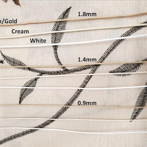 May include: Close-up of several cords in tan/gold, cream, and white, with varying thicknesses of 1.8mm, 1.4mm, and 0.9mm. The cords are displayed against a beige fabric with embroidered black leaf patterns. The image shows the different cord colors and sizes.