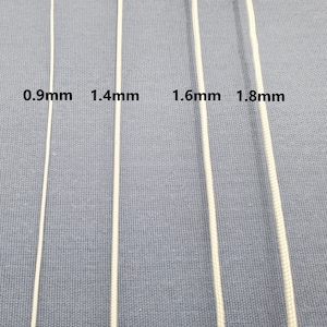 May include: Four white cords of varying thicknesses are displayed against a blue fabric background. The cords are labeled with their diameters: 0.9mm, 1.4mm, 1.6mm, and 1.8mm. The image is a close-up, showcasing the texture and size differences.