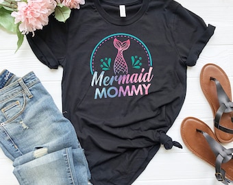 May include: Black t-shirt with a colorful "Mermaid Mommy" graphic. The design features a mermaid tail and the text in a gradient of pink, blue, and purple. The shirt is paired with blue jeans and brown sandals.