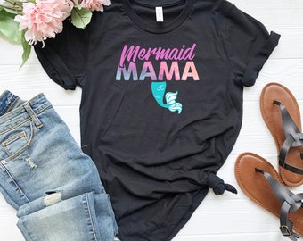 May include: Black t-shirt with the text "Mermaid MAMA" in pink and blue, and a turquoise mermaid tail graphic. The shirt is paired with blue jeans and brown sandals, with pink flowers in the background.