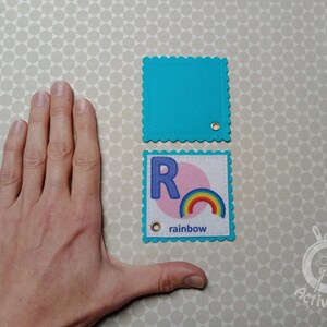 K&ouml;nnte beinhalten: Zwei Filzquadrate mit gewellten Kanten. Das obere Quadrat ist t&uuml;rkisblau. Das untere Quadrat ist t&uuml;rkisblau mit einem wei&szlig;en Kreis und dem Buchstaben "R" in blau. Der Text "rainbow" ist unter dem Kreis gedruckt.