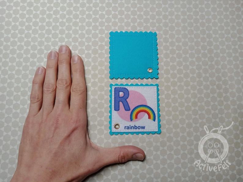 K&ouml;nnte beinhalten: Zwei Filzquadrate mit gewellten Kanten. Das obere Quadrat ist t&uuml;rkisblau. Das untere Quadrat ist t&uuml;rkisblau mit einem wei&szlig;en Kreis und dem Buchstaben "R" in blau. Der Text "rainbow" ist unter dem Kreis gedruckt.