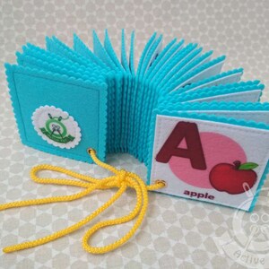 K&ouml;nnte beinhalten: Ein blaues Filz-Alphabetbuch mit einem gelben Schn&uuml;rsenkel. Das Buch ist auf den Buchstaben A ge&ouml;ffnet, der braun ist und eine rote Apfel-Illustration daneben hat. Der Text "apple" ist unter dem Apfel gedruckt.
