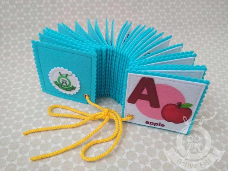 K&ouml;nnte beinhalten: Ein blaues Filz-Alphabetbuch mit einem gelben Schn&uuml;rsenkel. Das Buch ist auf den Buchstaben A ge&ouml;ffnet, der braun ist und eine rote Apfel-Illustration daneben hat. Der Text "apple" ist unter dem Apfel gedruckt.