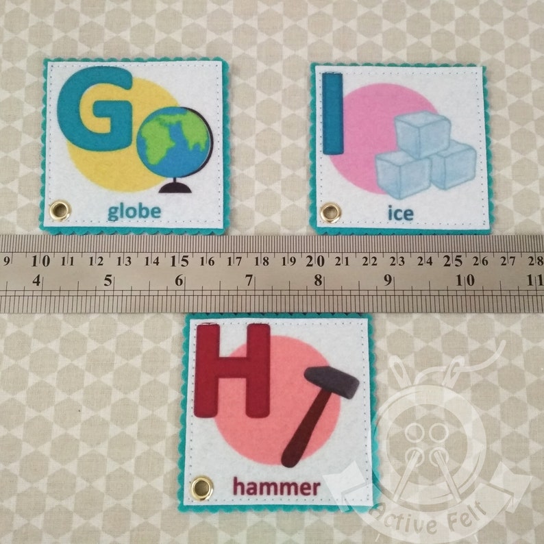 K&ouml;nnte beinhalten: Drei Filzquadrate mit einem Buchstaben des Alphabets, einem Bild und einem Wort auf jedem. Das erste Quadrat ist t&uuml;rkisfarben mit wei&szlig;em Hintergrund und dem Buchstaben G in T&uuml;rkis. Das Bild ist ein Globus und das Wort ist "Globus". Das zweite Quadrat ist t&uuml;rkisfarben mit wei&szlig;em Hintergrund und dem Buchstaben I in T&uuml;rkis. Das Bild sind drei Eisw&uuml;rfel und das Wort ist "Eis". Das dritte Quadrat ist t&uuml;rkisfarben mit wei&szlig;em Hintergrund und dem Buchstaben H in Rotbraun. Das Bild ist ein Hammer und das Wort ist "Hammer".