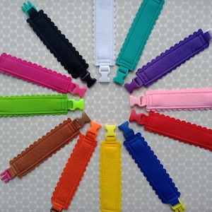 Op de afbeelding: Een set van 12 kleurrijke feltarmbanden met gegolfde randen en plastic sluitingen. De armbanden zijn in een cirkel gerangschikt, met de sluitingen naar buiten gericht.