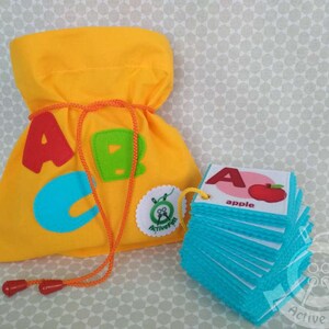 K&ouml;nnte beinhalten: Eine gelbe Filztasche mit den Buchstaben A, B und C darauf. Die Tasche hat einen Kordelverschluss. Es gibt einen Stapel blauer Filzkarten mit Buchstaben und Bildern darauf. Die oberste Karte hat den Buchstaben A und ein Bild eines Apfels mit dem Wort "Apfel" darunter.