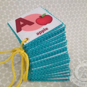 K&ouml;nnte beinhalten: Ein Stapel Filz-Alphabet-Karten mit einer roten Apfel-Illustration und dem Wort "apple" auf der obersten Karte. Die Karten haben einen t&uuml;rkisfarbenen gewellten Rand und sind mit einer gelben Schnur zusammengebunden.