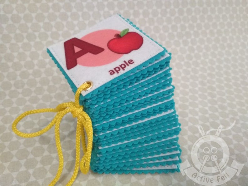 K&ouml;nnte beinhalten: Ein Stapel Filz-Alphabet-Karten mit einer roten Apfel-Illustration und dem Wort "apple" auf der obersten Karte. Die Karten haben einen t&uuml;rkisfarbenen gewellten Rand und sind mit einer gelben Schnur zusammengebunden.