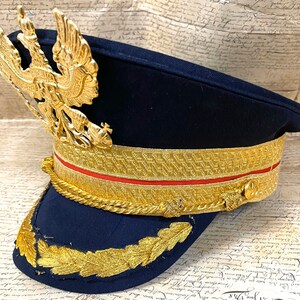 May include: A dark blue military style hat with a gold braid trim and a gold eagle emblem on the front.