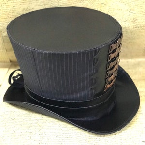 May include: A black top hat with a pinstripe pattern and a decorative band of metal accents. The hat has a wide brim and a tall crown.