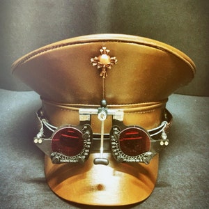 May include: A brown leather military-style cap with a skull and crossbones design on the top. The cap has a pair of black goggles attached to the front.