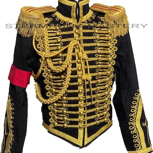 Può includere: Giacca nera in stile militare con decorazioni dorate. Presenta bottoni dorati, finiture, spalline e corda decorativa. Una fascia rossa è sulla manica sinistra. La giacca ha un colletto alto.