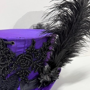 Pu&ograve; includere: Un cappello viola con abbellimenti neri e una grande piuma nera. Il cappello presenta intricate perline nere, pizzo e un piccolo fiore nero. Un velo di rete nero &egrave; attaccato al cappello.