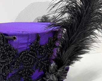 May include: A purple hat with black embellishments and a large black feather. The hat features intricate black beadwork, lace, and a small black flower. A black net veil is attached to the hat.