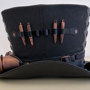 May include: A black top hat with a black leather band and a copper-colored steampunk-style embellishment. The hat has a black fabric band with five copper-colored bullets attached.