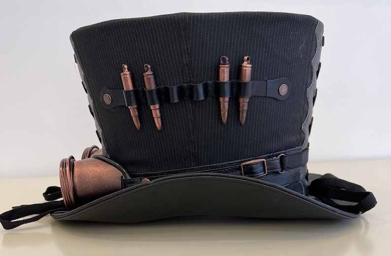 May include: A black top hat with a black leather band and a copper-colored steampunk-style embellishment. The hat has a black fabric band with five copper-colored bullets attached.