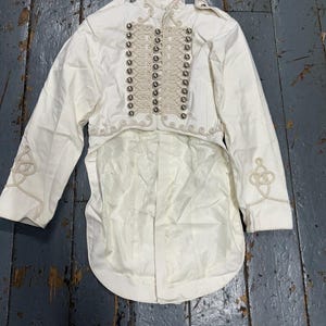 May include: Cream military-style jacket with silver buttons and decorative trim. The jacket has long sleeves and a tailcoat design. The garment features decorative embroidery on the sleeves.