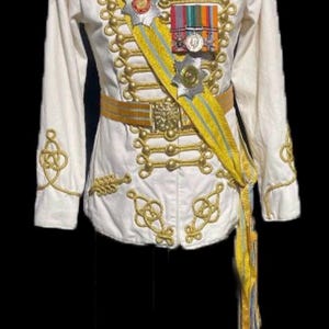 Può includere: Una giacca militare bianca con rifiniture e bottoni dorati. La giacca ha una fascia con una stella e medaglie. La giacca è su un manichino.