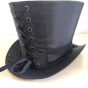 Op de afbeelding: Zwarte hoge hoed met een brede rand en een satijnen afwerking. De hoed heeft een veter aan de zijkant met bronzen oogjes en een zwarte lintband. Een klassiek accessoire voor cosplay of thema-evenementen.