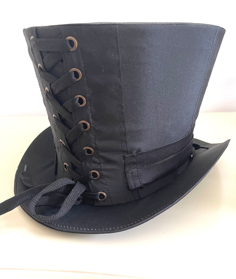 Op de afbeelding: Zwarte hoge hoed met een brede rand en een satijnen afwerking. De hoed heeft een veter aan de zijkant met bronzen oogjes en een zwarte lintband. Een klassiek accessoire voor cosplay of thema-evenementen.