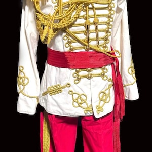 Può includere: Un costume in stile militare con una giacca bianca con finiture dorate, spalline e ricami decorativi. L'outfit include pantaloni rossi con strisce dorate e una fascia rossa. Per teatro o cosplay.